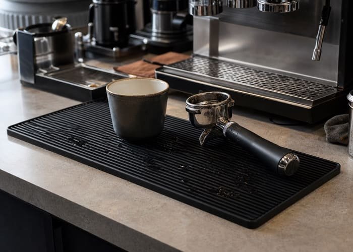 A sleek, minimalist black silicone bar mat on a polished marble coffee counter, with a steaming espresso cup on it.