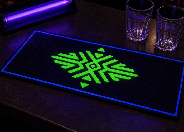 A UV fluorescent custom bar mat on a dark bar top, with a glowing green logo under a blacklight